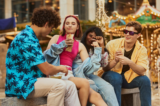 Young people eating wraps and chatting near amusement ride. Concept of youth culture visuals, event branding, modern food promotion, emotional storytelling.