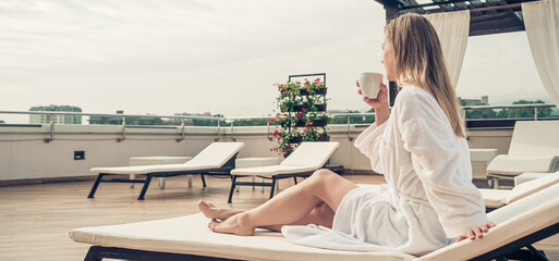 Woman in white bathrobe lying on sofa and relaxing with orange cocktail at the roof.