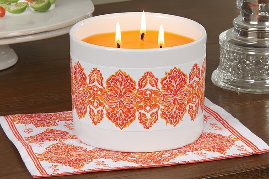 Round, white ceramic candle with orange-red floral design.  Three flickering flames.  On a wooden surface, with a patterned napkin.  Details of other items visible in the background