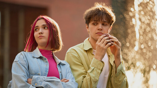 Young girl disappointed while boy eats suspicious burger near fountain. Concept of food miscommunication, awkward street moments, youth storytelling for modern campaigns.
