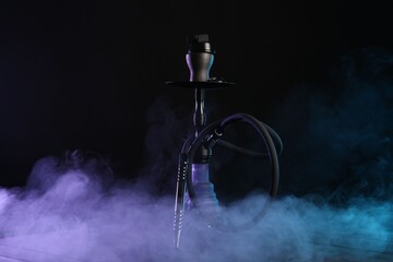 Hookah on wooden table with smoke in color lights against black background