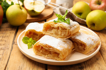 Pieces of tasty apple strudel with powdered sugar, mint and fruits on wooden table, closeup