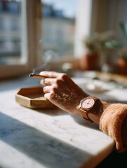 Close-up of a hand holding a cigarette, watch visible, on a sunlit coffee table, creating a peaceful morning vibe.