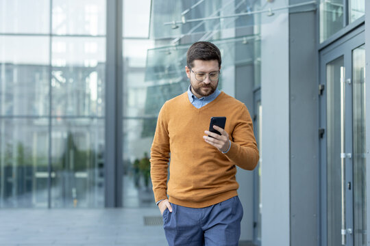 Serious young man walking down the street near an office building, holding his hand in his pocket and using his mobile phone