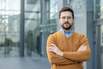 Obraz premium Close-up portrait of a serious young confident businessman standing on the street near his office, arms crossed on his chest and looking at the camera.