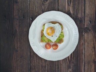 Top view of Heart shaped fried egg with pepper on smashed avocado and tomato on a white plate on a wooden background