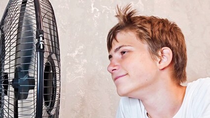 Teenager boy suffering from the heat at home, he is sitting on the couch next to an electric fan....