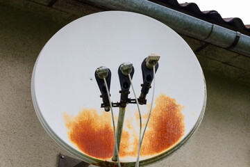 An old grey satellite dish, mounted on a building wall, shows significant signs of rust. This image...