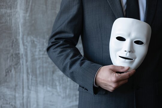 Businessman in suit holding white mask, hidden identity, deception, corporate fraud and disguise concept.