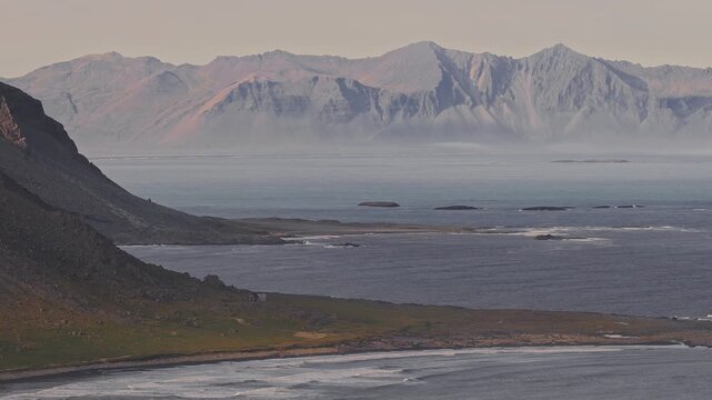 A slow sweeping aerial pan of Hvalnes cliffs, rugged terrain, scattered islets, and mist hovering near the coastline in Iceland.