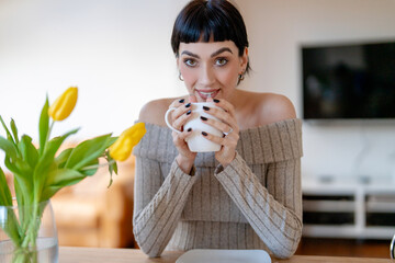 Woman enjoying coffee while sitting at a wooden table adorned with yellow tulips during a cozy...