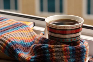 A cup of coffee surrounded by a warm knitted scarf against the backdrop of city windows. Coffee and a scarf on the balcony windowsill. The onset of autumn and winter.