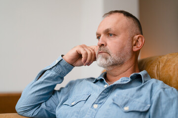 Man in casual shirt thoughtfully sitting on a couch in a cozy indoor setting during a quiet...