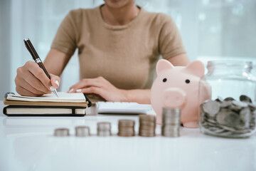 An Asian woman is writing in a notebook and stacking coins and a piggy bank, which symbolizes...