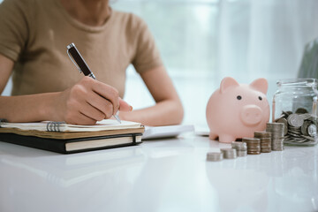 An Asian woman is writing in a notebook and stacking coins and a piggy bank, which symbolizes...