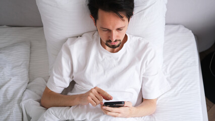 Young brunette man lying in bed looking at his phone early in the morning in a bright cozy bedroom