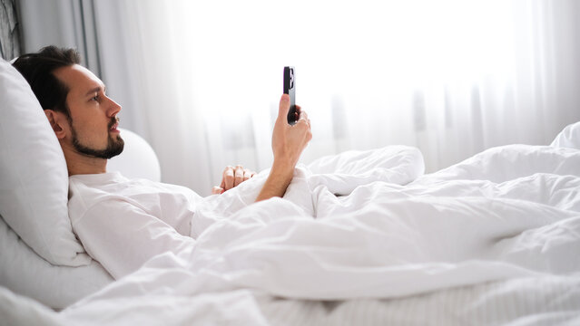 Young brunette man lying in bed looking at his phone early in the morning in a bright cozy bedroom