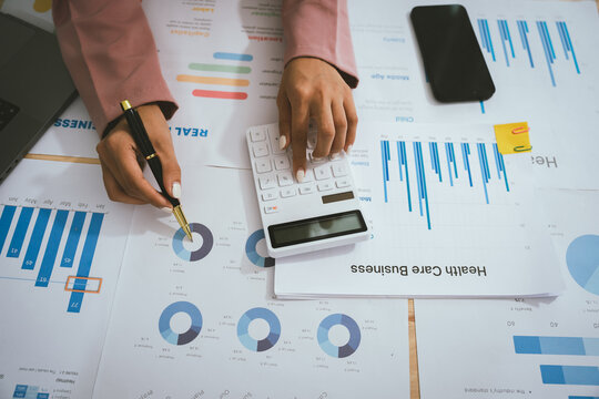 Businesswoman analyzing financial data using calculator, holding documents surrounded by charts, graphs and laptop, showing professionalism, business planning.