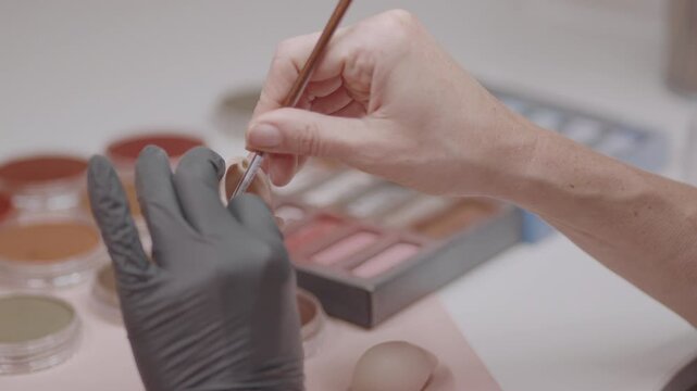 Artist painting small silicone doll held in gloved hand using fine brush, surrounded by makeup tools, showing precision in realistic detailing process