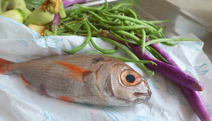 Fresh fish on paper in kitchen before cooking on table. Vegetables in background. Food and organic seafood.