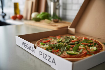 Freshly delivered vegetable pizza in box labeled "vegan pizza" on kitchen counter. Healthy plant-based fast food option at home