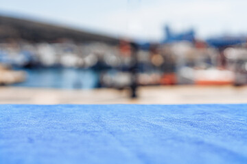 A blurred view of a harbor with boats in the background. The foreground features a blue tablecloth. The scene conveys a relaxed maritime atmosphere.