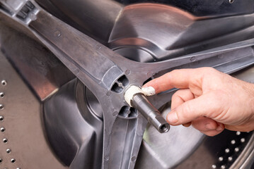 Lubricating the washing machine shaft during repair. A hand applies white grease to the metal shaft of a washing machine drum, an important step in assembly or servicing