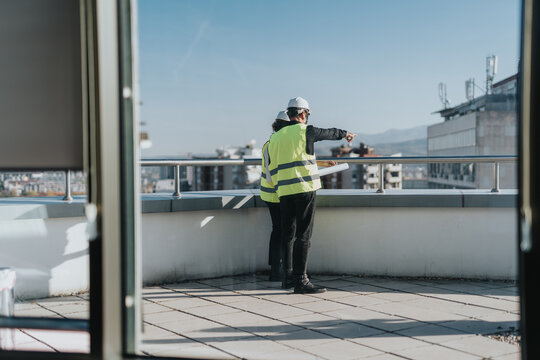 Two construction engineers wearing safety vests and helmets discuss building plans on a rooftop, with a cityscape in the background. They appear focused on the progress of the project. - Powered by Adobe
