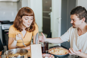 Two young friends share a joyful moment while eating pasta and drinking beverages in a cozy...