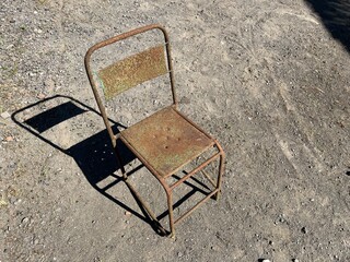Old rusty metal chair standing alone on dry gravel ground in sunlight, showing weathered texture and aged condition.