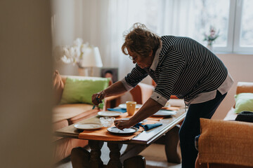 A senior woman setting up the dining table in a warmly lit home, displaying attentiveness and care in arranging the space for a meal in a home-like environment.