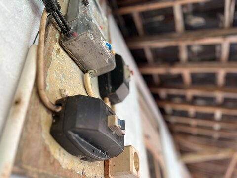 Close-up of old electric meters and circuit boxes with exposed wires mounted on a worn wall under a wooden roof, showing aged infrastructure and utility equipment.