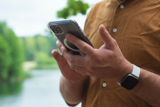 Close-up of a man using a smartphone with a transparent case, standing outdoors near a lake. Concept modern digital lifestyle, mobile connectivity on the go in a natural environment.