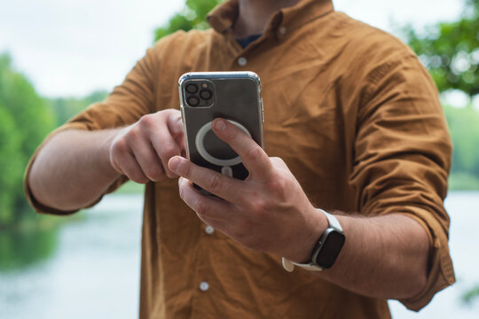 Close-up of a man using a smartphone with a transparent case, standing outdoors near a lake. Concept modern digital lifestyle, mobile connectivity on the go in a natural environment. - Powered by Adobe