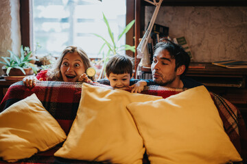 A cheerful family smiling playfully behind a cozy sofa with pillows, creating a warm and joyful atmosphere indoors, surrounded by books and plants.