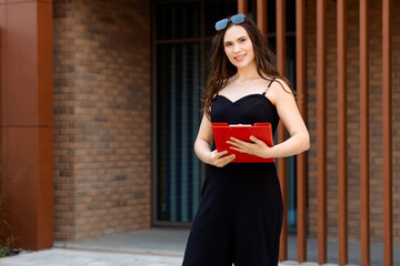 Young woman in black dress holding red tablet, smiling against brick wall, radiating confidence and...