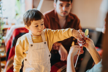 A young boy reaches out to take a lollipop from an adult during a warm family gathering in a living room. A male adult observes the interaction, adding to the familial feel.