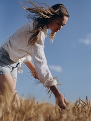 Young woman stands and collects wheat earls in golden wheat field under blue sky.