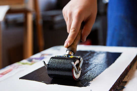 A closeup of an artist hand using a brayer to apply black ink onto a linocut surface in a linocut printmaking process. 

