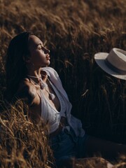 Young woman relaxes sitting with closed eyes near straw hat on field among ripe ears of wheat.