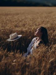 Young woman relaxes sitting with closed eyes near straw hat on field among ripe ears of wheat.