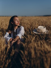 Young woman relaxes sitting with closed eyes near straw hat on field among ripe ears of wheat.