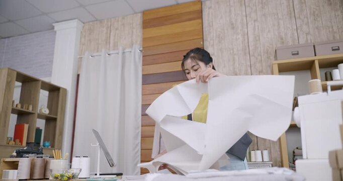 Asian female dressmaker unfolding paper prototype and reviewing alignment for garment cutting in tailoring studio with handmade fashion tools and clothing items arranged in workspace interior