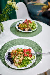 Fresh salad with vibrant vegetables on white plate, set on round table with green placemat. Soft indoor lighting enhances inviting ambiance alongside lush greenery centerpiece