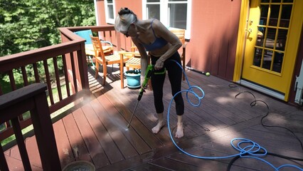 A mature woman utilizes a pressure washer to clean a wooden deck in her backyard, effectively removing dirt and stains from the surface.