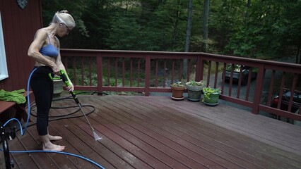 A mature woman uses a pressure washer to clean dirt off her wooden deck in a tranquil backyard setting. She focuses on the task while barefoot.