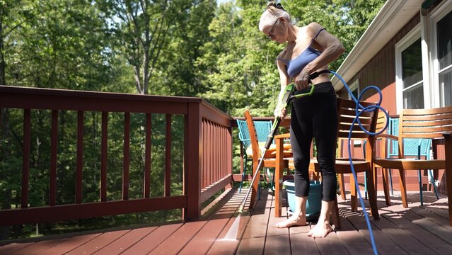 A mature woman is using a pressure washer to clean her wooden deck, ensuring it is free of dirt and grime while working in a bright garden setting.