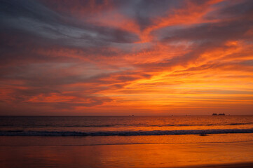 A vibrant sunset paints the sky with fiery hues of orange and red, reflecting on the tranquil ocean waters and sandy beach below, creating a breathtaking scene of natural beauty.