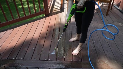 A mature woman is pressure washing her wooden deck in the backyard, removing dirt and stains to enhance the outdoor space for home improvement.