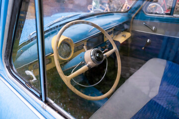 A close-up through the window of a vintage car's interior, focusing on the steering wheel, dashboard, and blue and white patterned seat, evoking a sense of nostalgia and retro style.